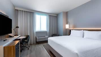 A hotel room with a bed, desk, chair, TV, air conditioning unit, and a window with curtains.
