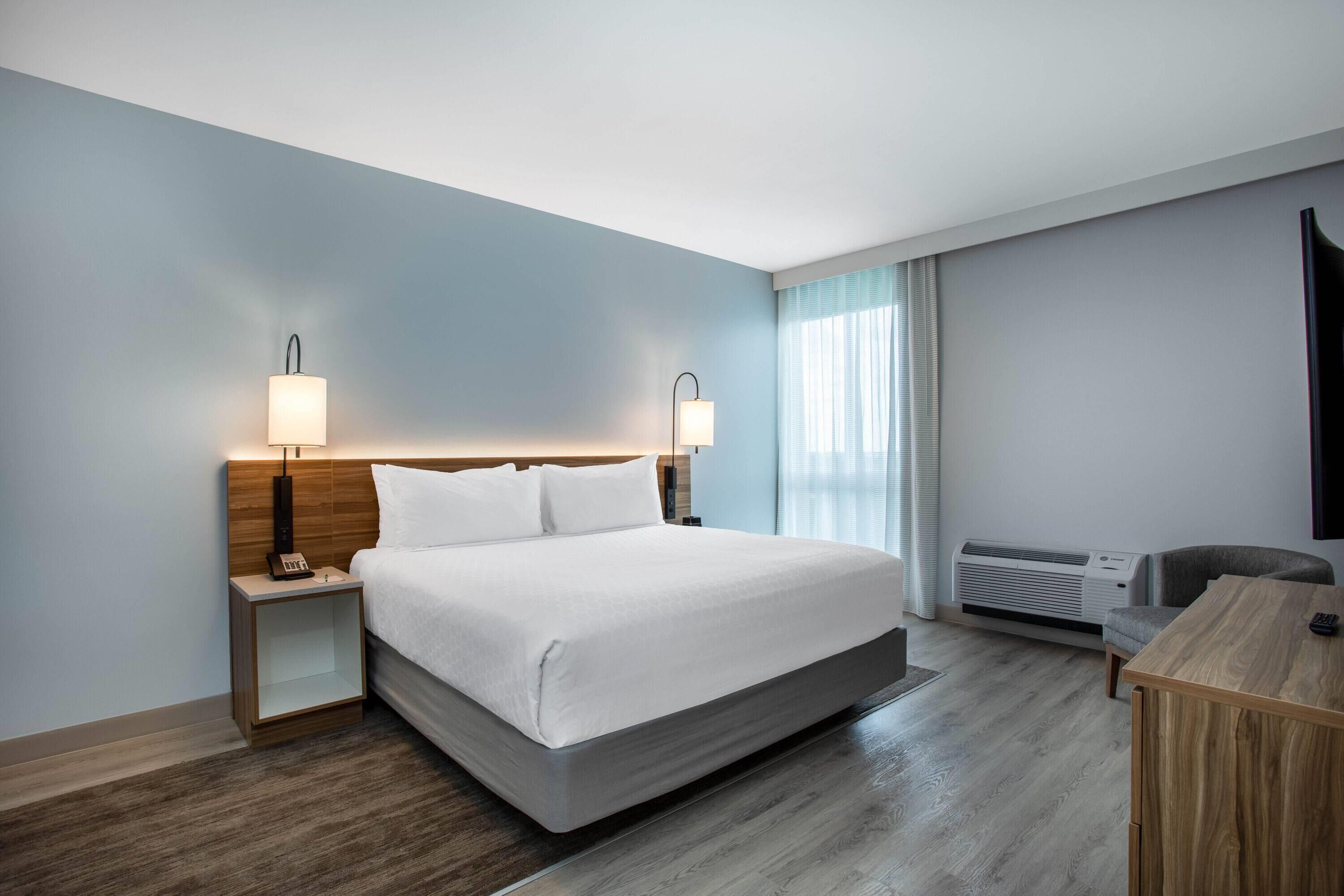 Une chambre d’hôtel avec un lit, des lampes de chevet, un bureau et une fenêtre avec des rideaux.