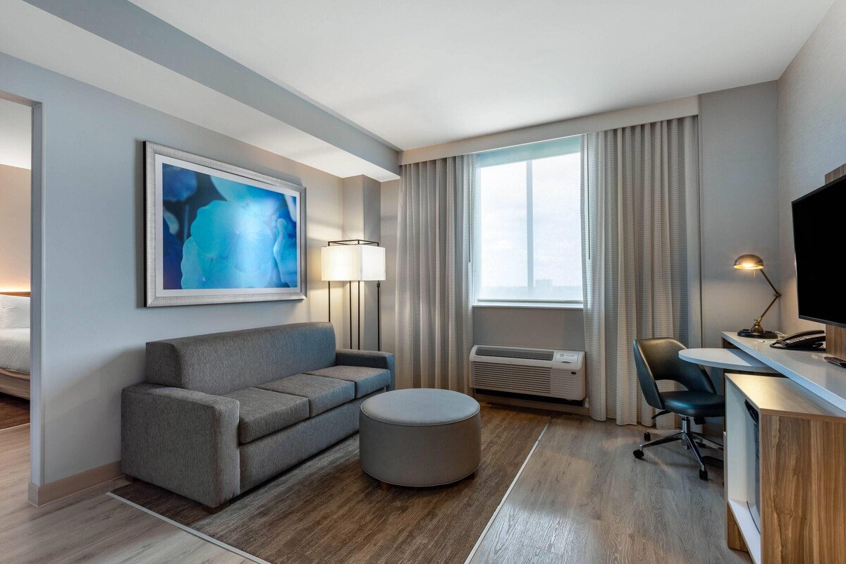 Una habitación de hotel moderna con un sofá gris, un otoman redond, un escritorio con una silla, un televisor de pantalla plana y un cuadro abstracto grande en la pared.