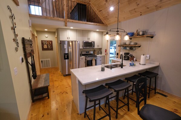 A modern kitchen with a large island, bar stools, stainless steel appliances, and wooden flooring. - Silver Lake Retreat - Beach Access + Dunes (Mears)