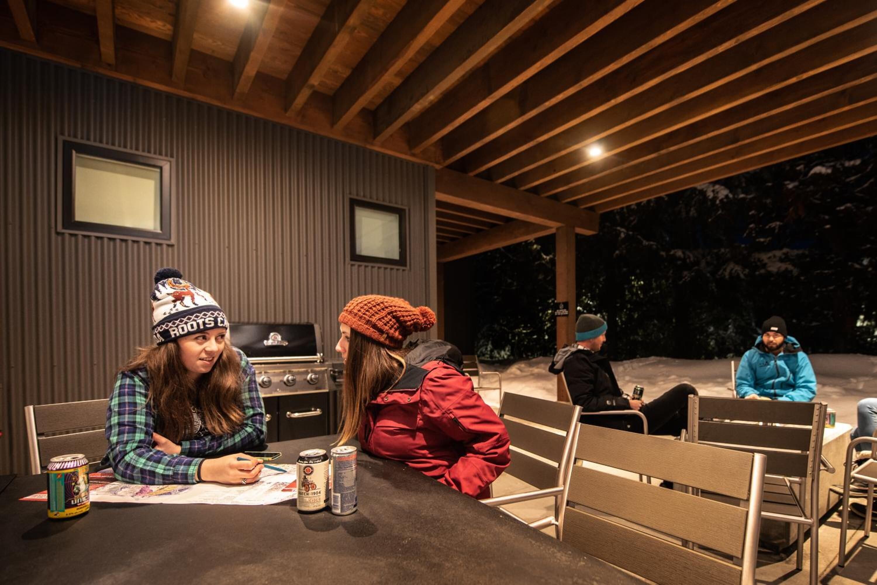 Uma área de refeições ao ar livre com pérgola de madeira, cadeiras de metal e uma mesa com comida e bebidas.