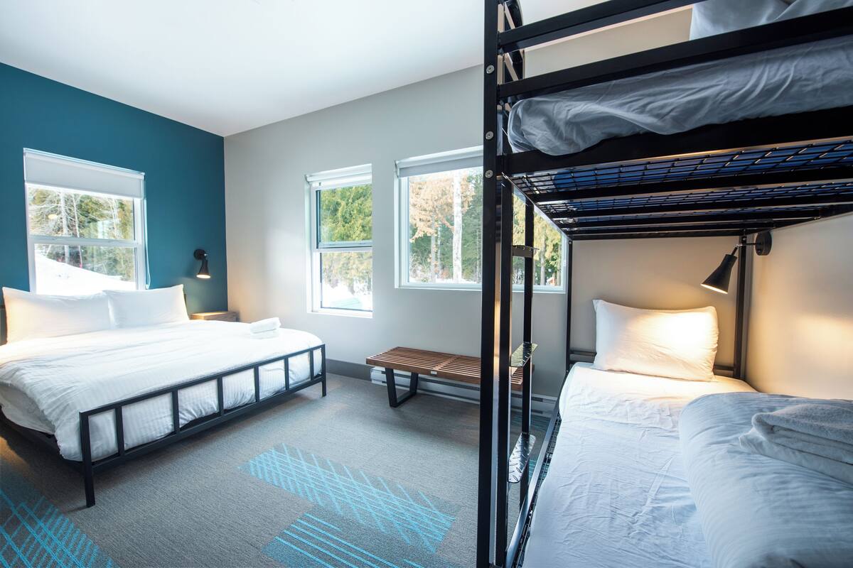 Una habitación de hotel moderna con una litera, una cama individual, un banco y ventanas amplias que dejan entrar luz natural.