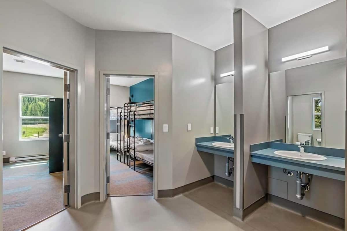 Un baño moderno con un espejo grande, dos lavamanos y una cama litera visible a través de una puerta abierta.