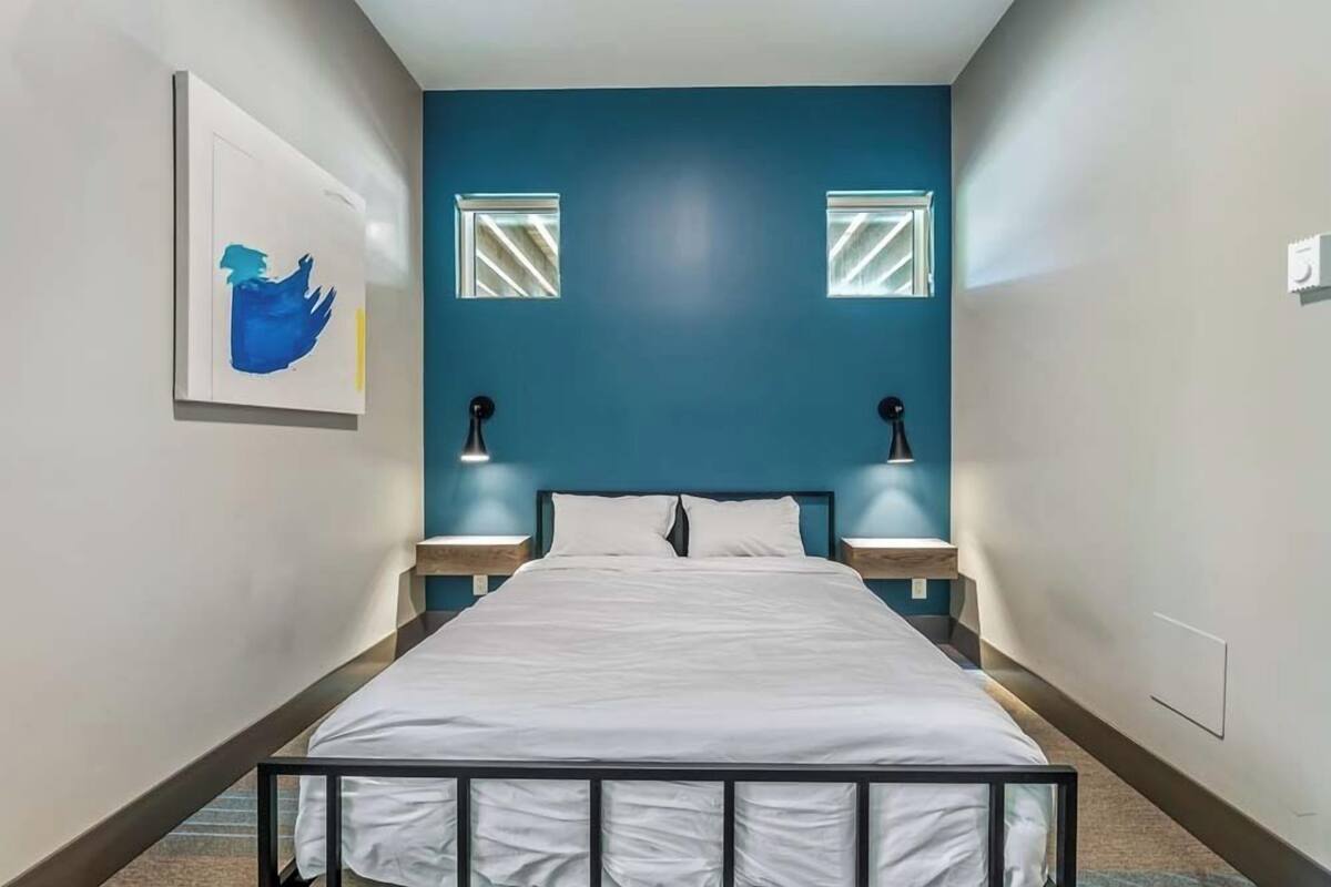 Un dormitorio con una cama, dos lámparas de noche, una obra de arte enmarcada en la pared y dos ventanas con persianas.