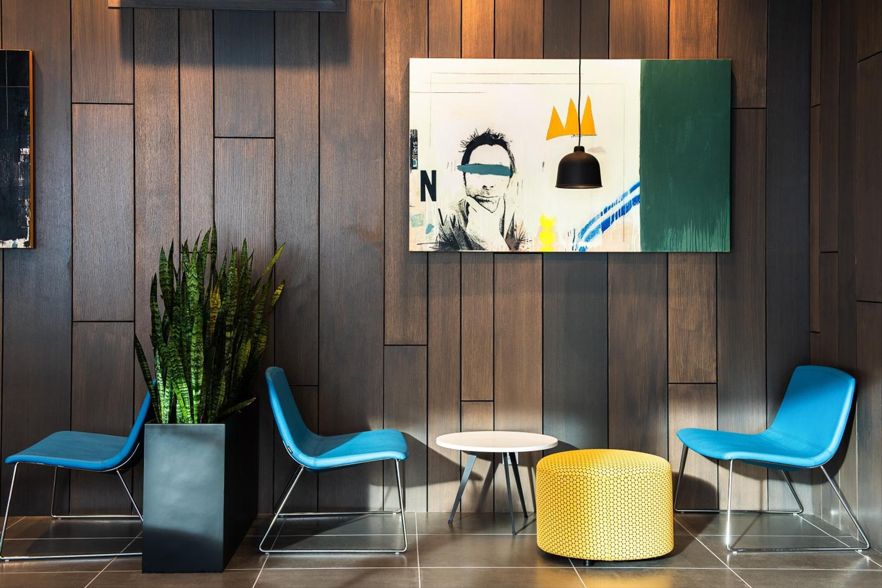 Een moderne lobby met houten paneelwanden, twee blauwe stoelen, een witte tafel, een geel poefje en een groot abstract schilderij.