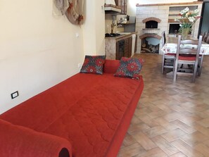 A red sofa with patterned cushions, a wooden dining table with chairs, a kitchen area with a stove and sink, and a fireplace.