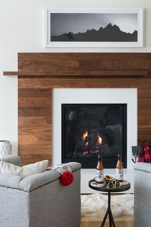 A living room with a fireplace, a sofa, a coffee table with bottles and glasses, and a framed picture above the fireplace.