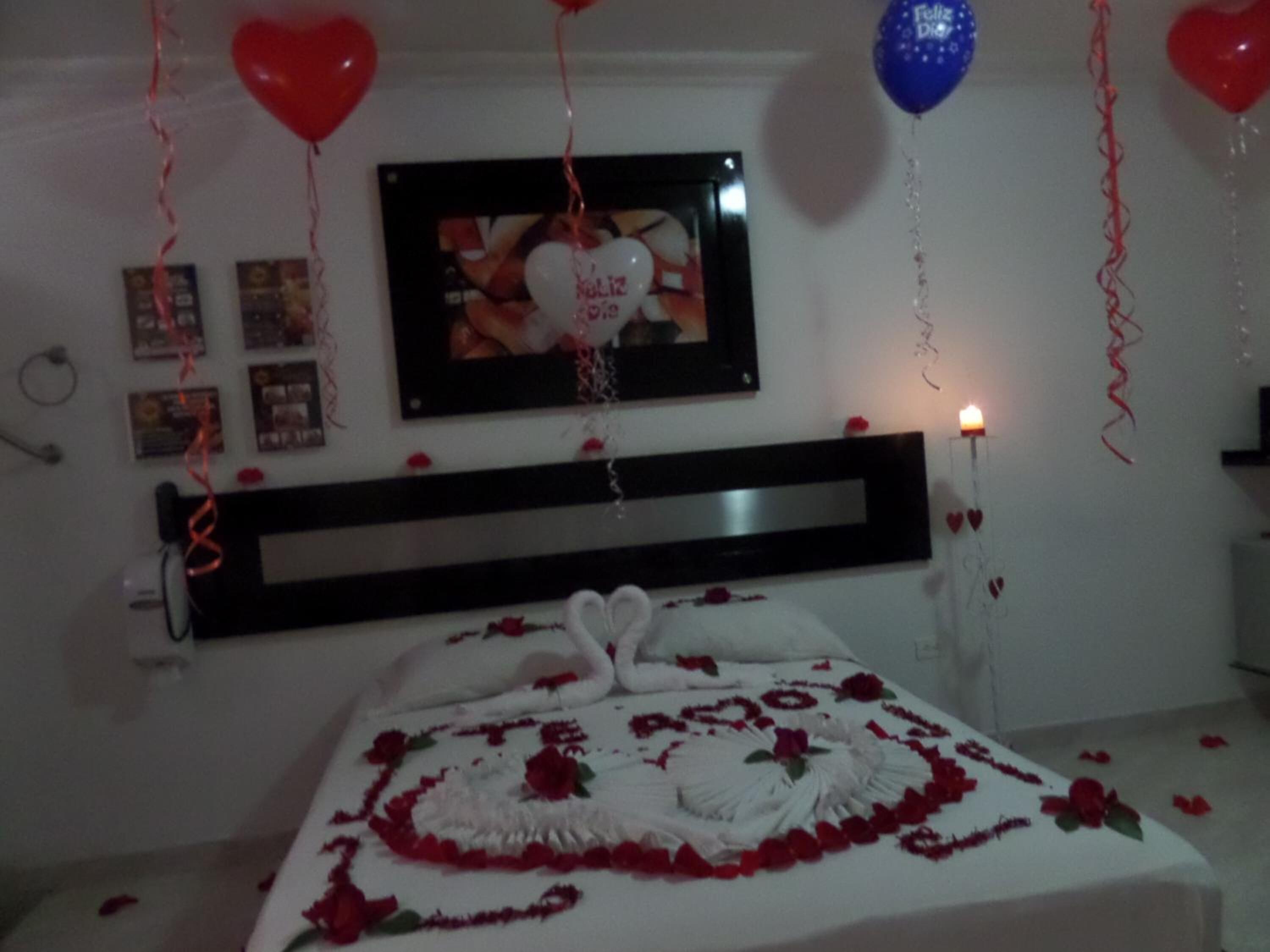 A bedroom with a bed adorned with rose petals and a heart-shaped decoration, a framed photo on the wall, and red and blue balloons.