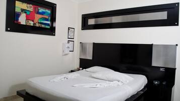 A modern hotel room with a black headboard, a bed with white linens, a framed abstract painting, a wall-mounted air purifier, and a small bedside table with a lamp.
