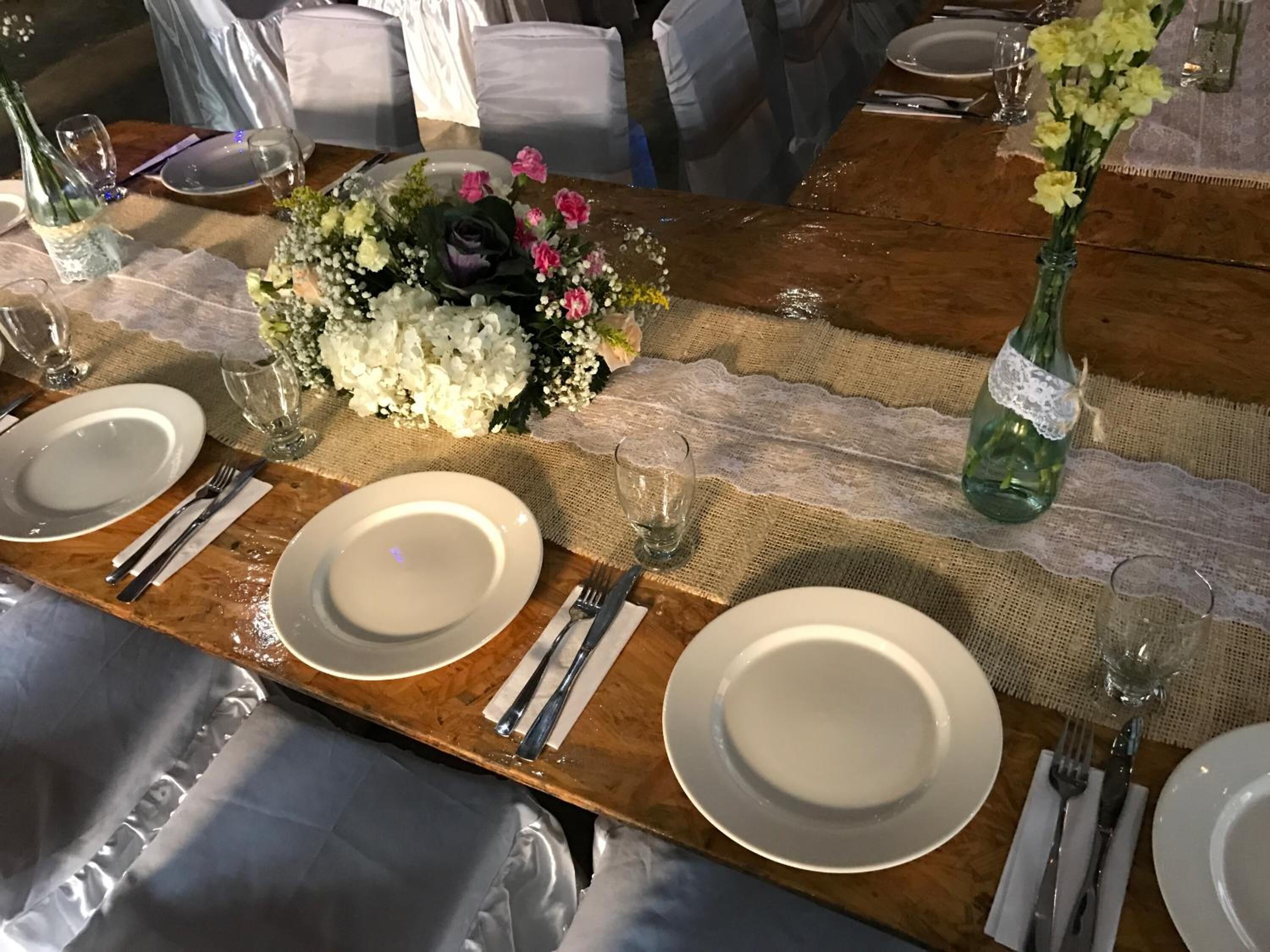 Una mesa de comedor lista para una comida, con platos blancos, cubiertos y un centro de mesa con flores.
