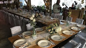 A long wooden table set with white plates, silverware, and glassware, surrounded by chairs with white covers. - Torettos (Yumbo)