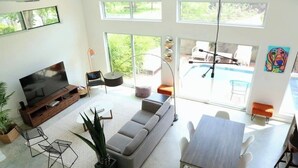 En moderne stue med TV, sofa, spisebord, stole og et stort vindue med udsigt til en pool.