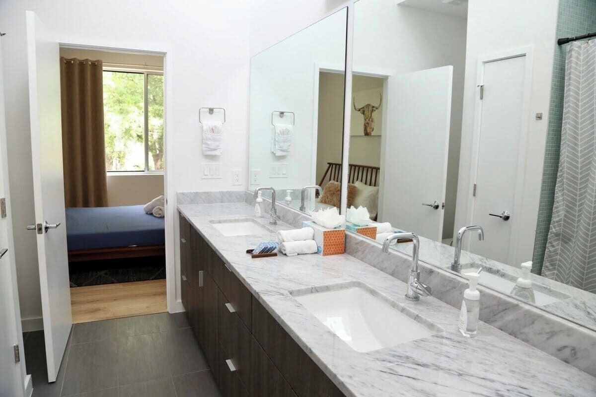 A bathroom with a large mirror, two sinks, a shower, and a bed with a view of greenery.