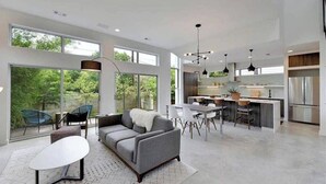 En rummelig opholdsstue med sofa, spisebord og stole, et køkken med moderne hårde hvidevarer, og store vinduer med udsigt til grønne områder.