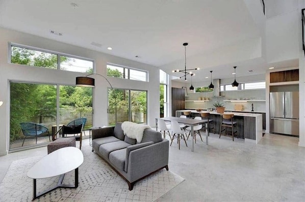 A spacious living area with a sofa, dining table, and chairs, a kitchen with modern appliances, and large windows offering a view of greenery.