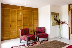 Una habitación con un armario de madera grande, dos sillones estampados, una mesita redonda pequeña, una alfombra grande y un cuadro en la pared.