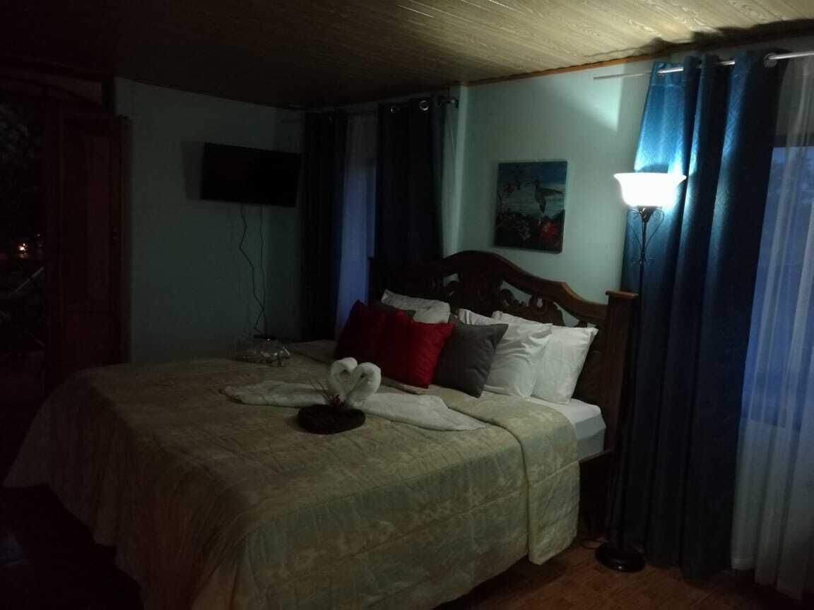 Habitación de hotel tenuemente iluminada con una cama, un televisor de pantalla plana montado en la pared y una lámpara en la mesita de noche.