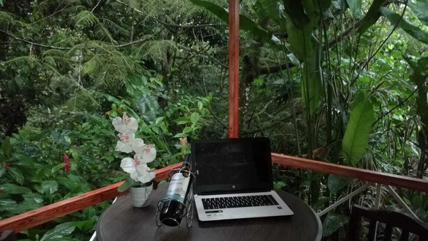 A laptop on a round table with a white flower vase and a thermos.