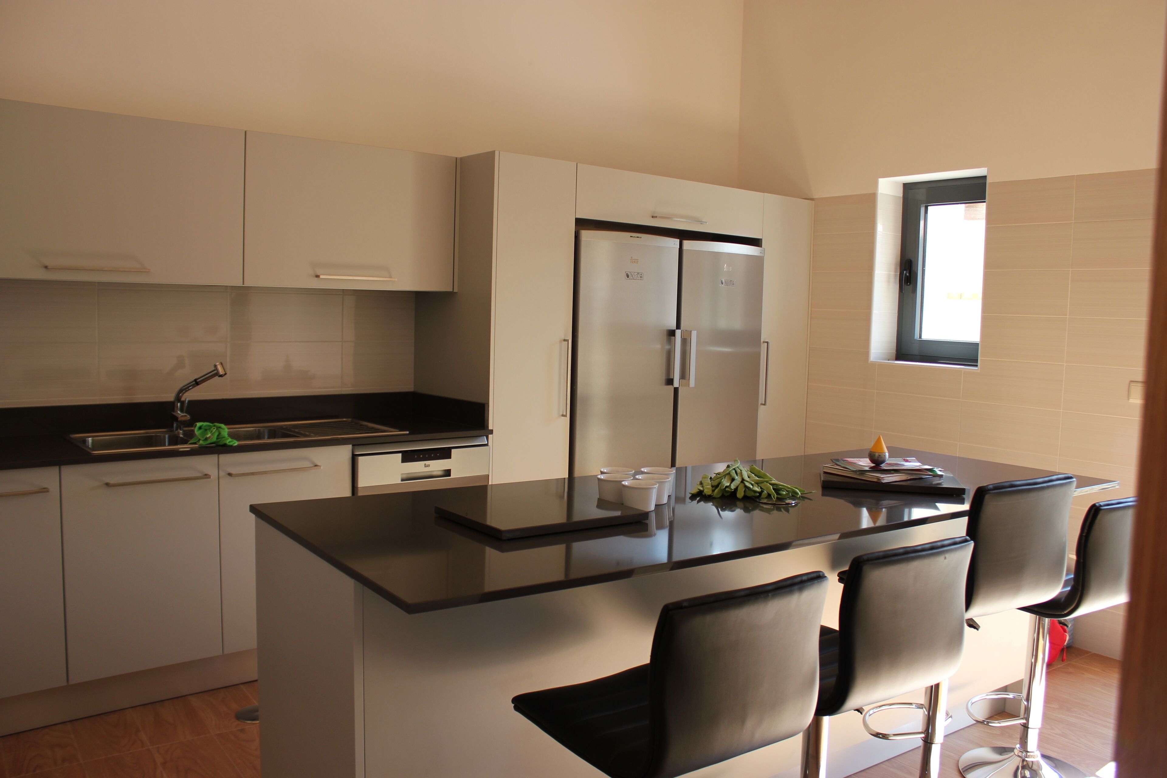 Een moderne keuken met witte kasten, een zwart aanrecht en roestvrijstalen apparatuur. Boven de gootsteen is een klein raam en er is een bar met stoelen.