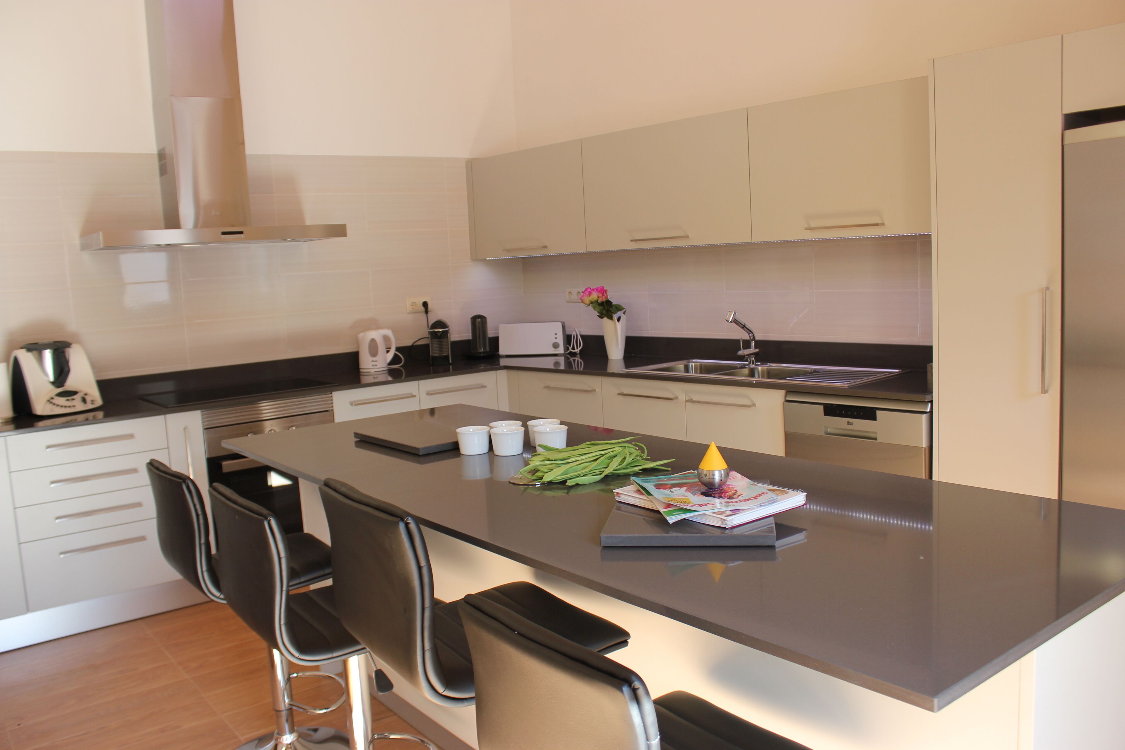 A modern kitchen with a central island, black bar stools, a built-in oven, and a stainless steel refrigerator.