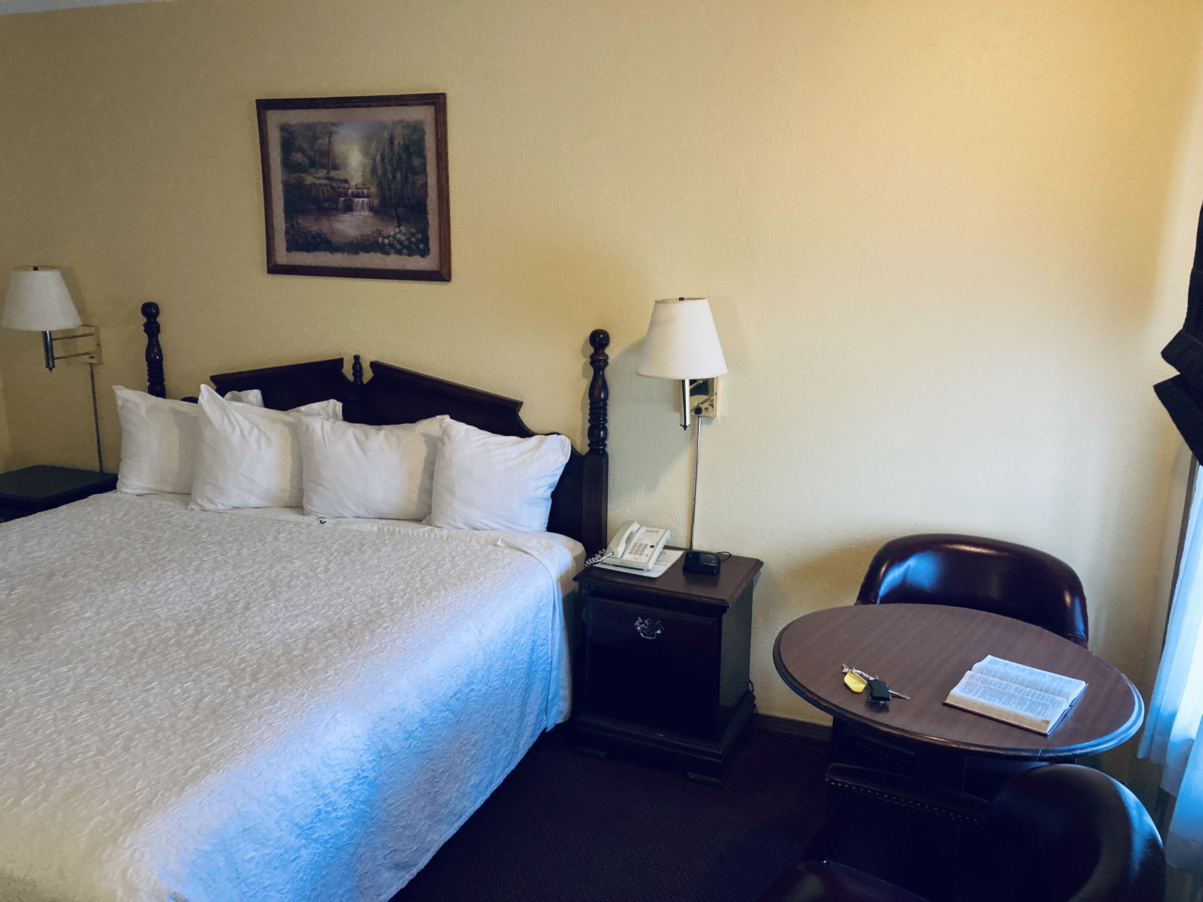 A hotel room with a large bed, two bedside tables with lamps, a small table with a chair, and a painting on the wall.