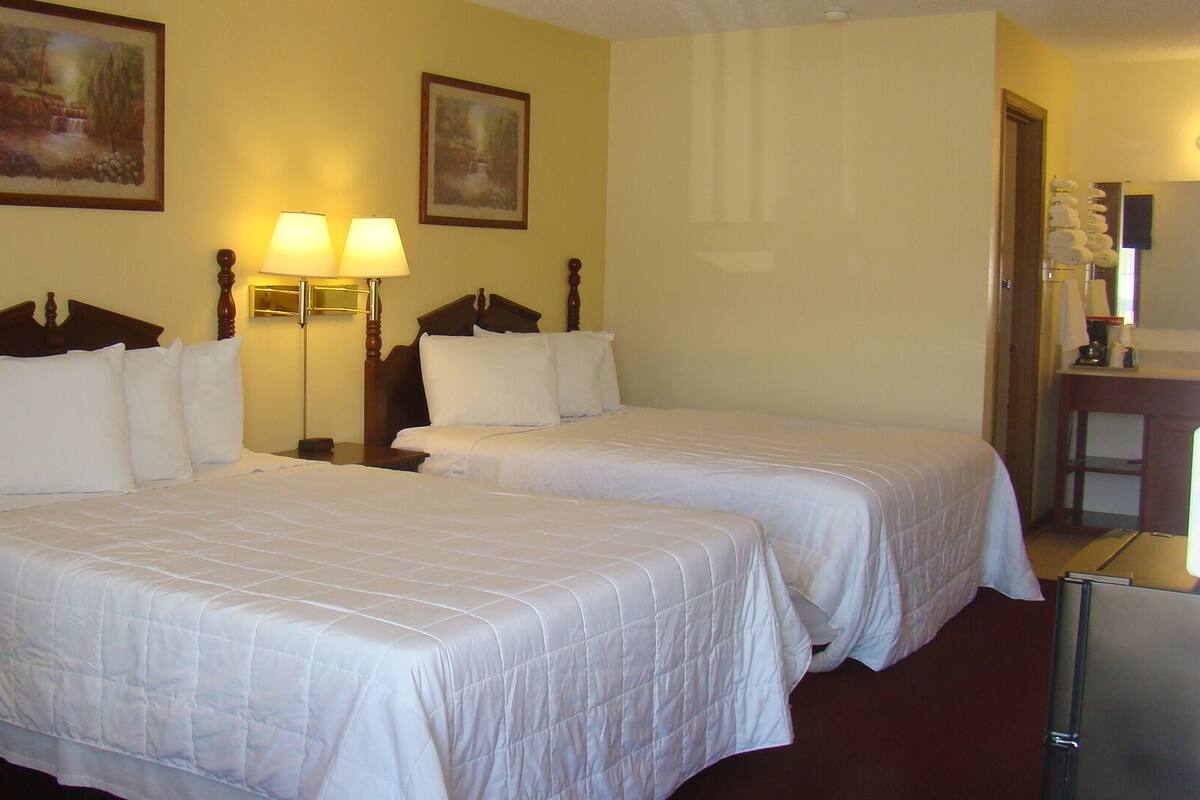 A hotel room with two beds, each with white bedding and wooden headboards. There are two wall-mounted lamps and two framed paintings above the beds. A small table with a lamp and a refrigerator are visible in the corner.