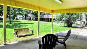 Un porche cubierto con una hamaca, dos sillas y vistas a un jardín con césped y casas.