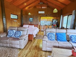 Interior acogedor de una cabaña con dos sofás, una cama, piso de madera y una zona de cocina.