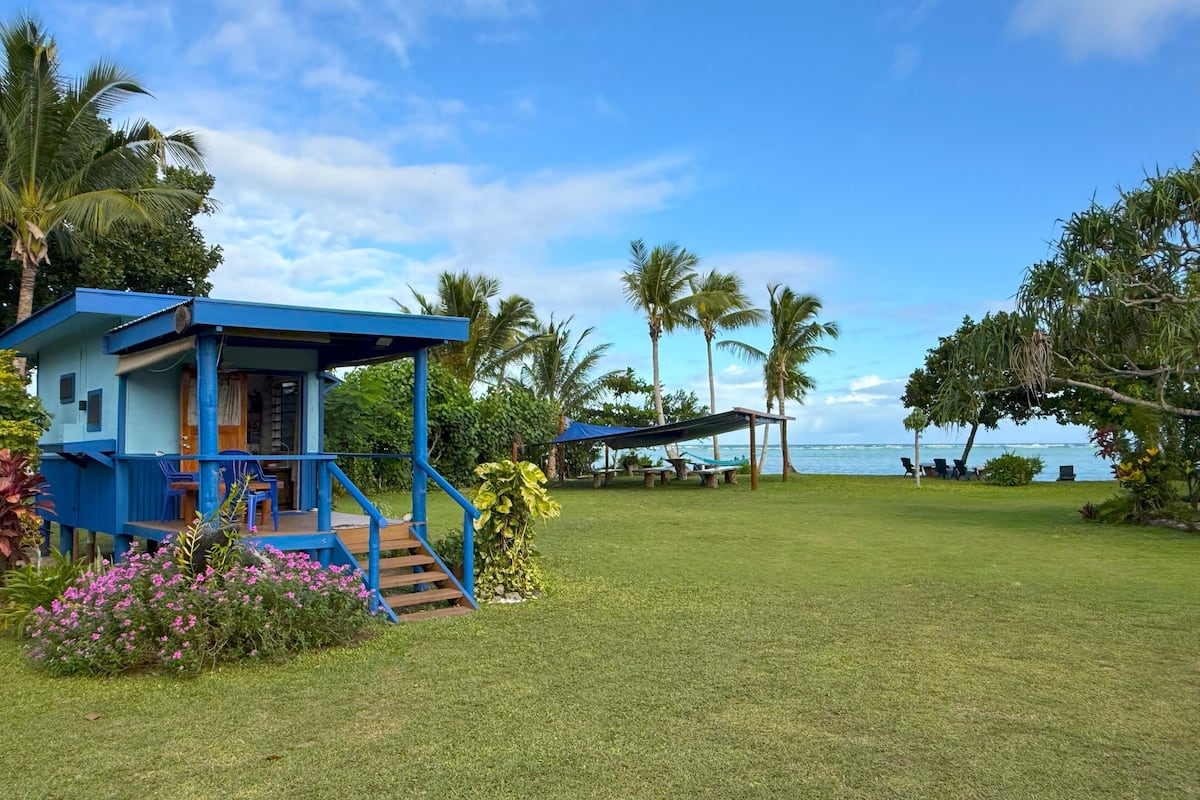 Een blauw gebouw met een veranda en een terras, omringd door gras en palmbomen, met uitzicht op een waterpartij.