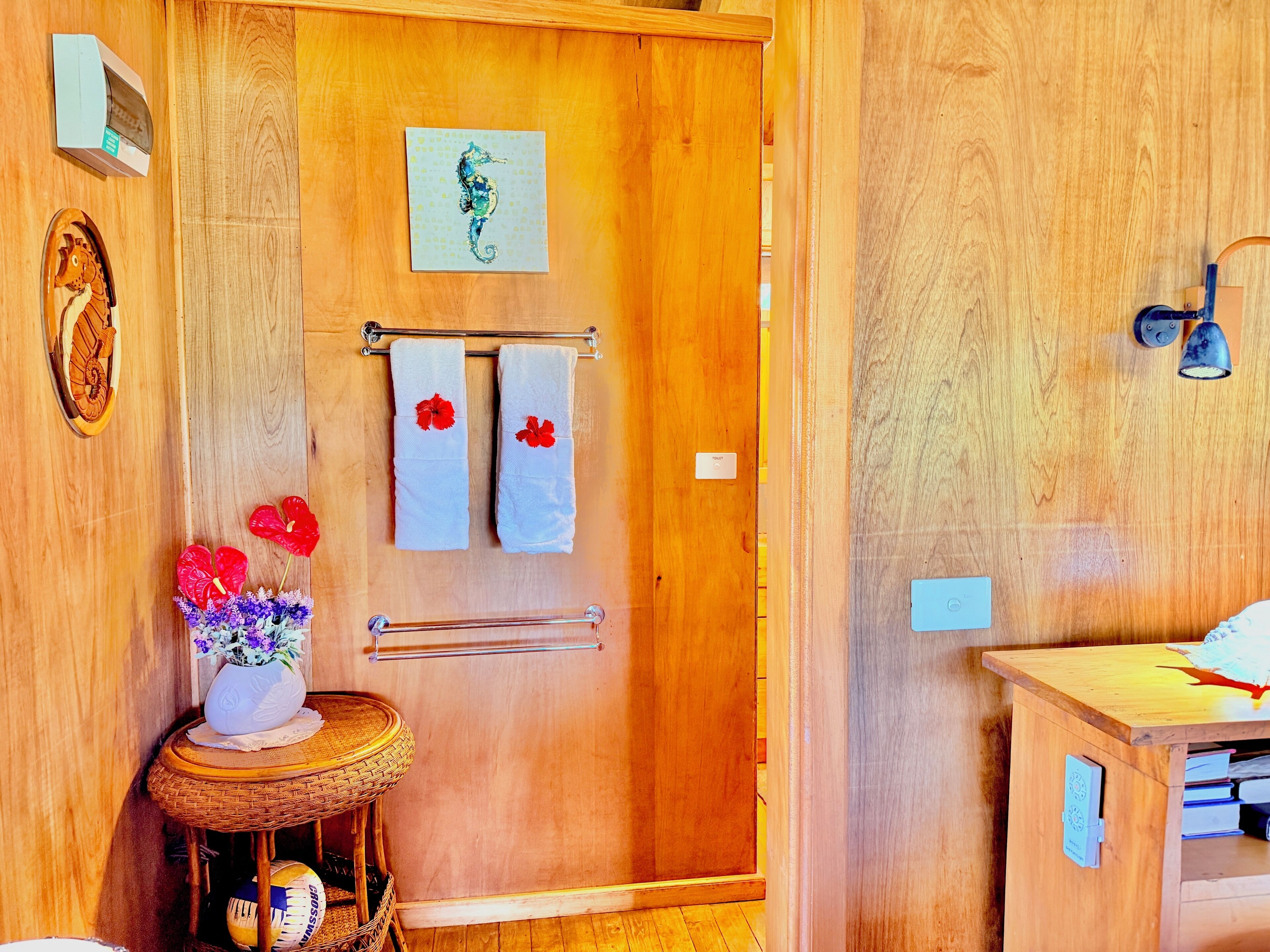 A wooden bathroom with a shower, a towel rack with two towels, a seahorse artwork, a wooden door, a small table with a vase, and a wall-mounted light fixture.