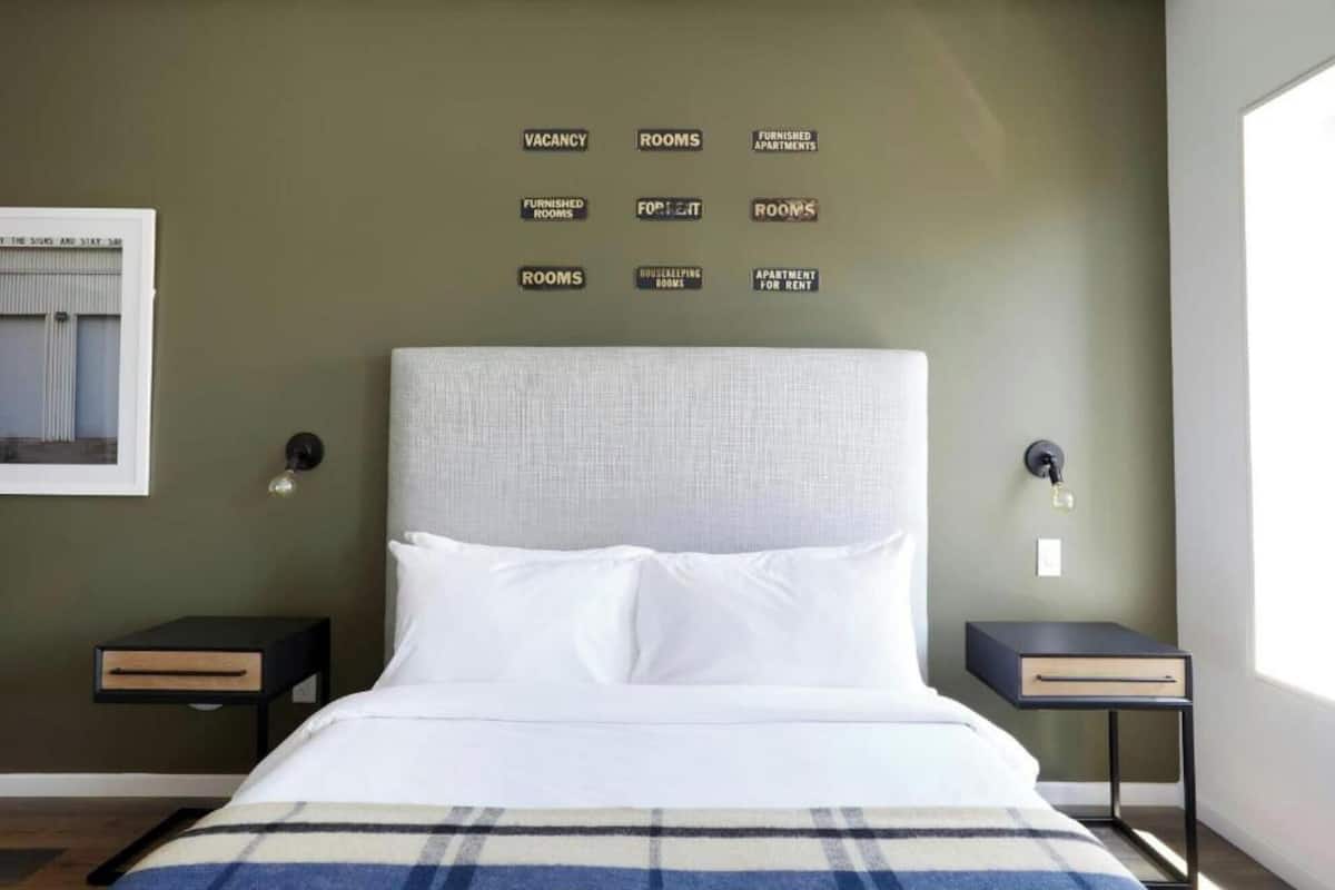 A bedroom with a bed, two bedside tables, a wall with multiple signs, and a window with curtains.