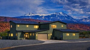 A two-story house with a garage, set against a mountainous backdrop. - Exclusive Retreats White Rim 3369- Luxury Moab Retreat with Private Hot Tub (Moab)
