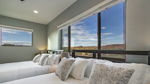 A hotel room with a large bed, multiple pillows, a view of a desert landscape, and a window with a view of a building. - Exclusive Retreats White Rim 3369- Luxury Moab Retreat with Private Hot Tub (Moab)