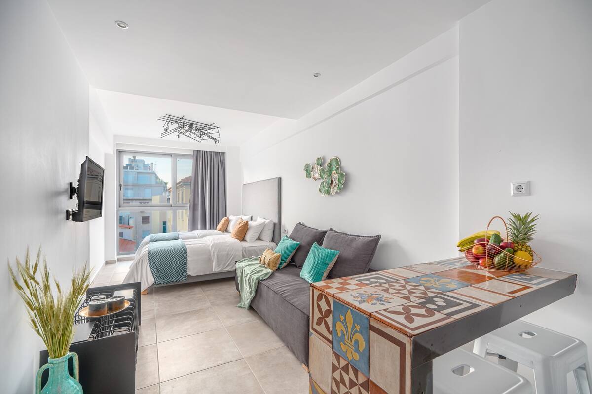 Una habitación moderna y bien iluminada con cama, sofá, pequeña cocina y vistas a edificios.