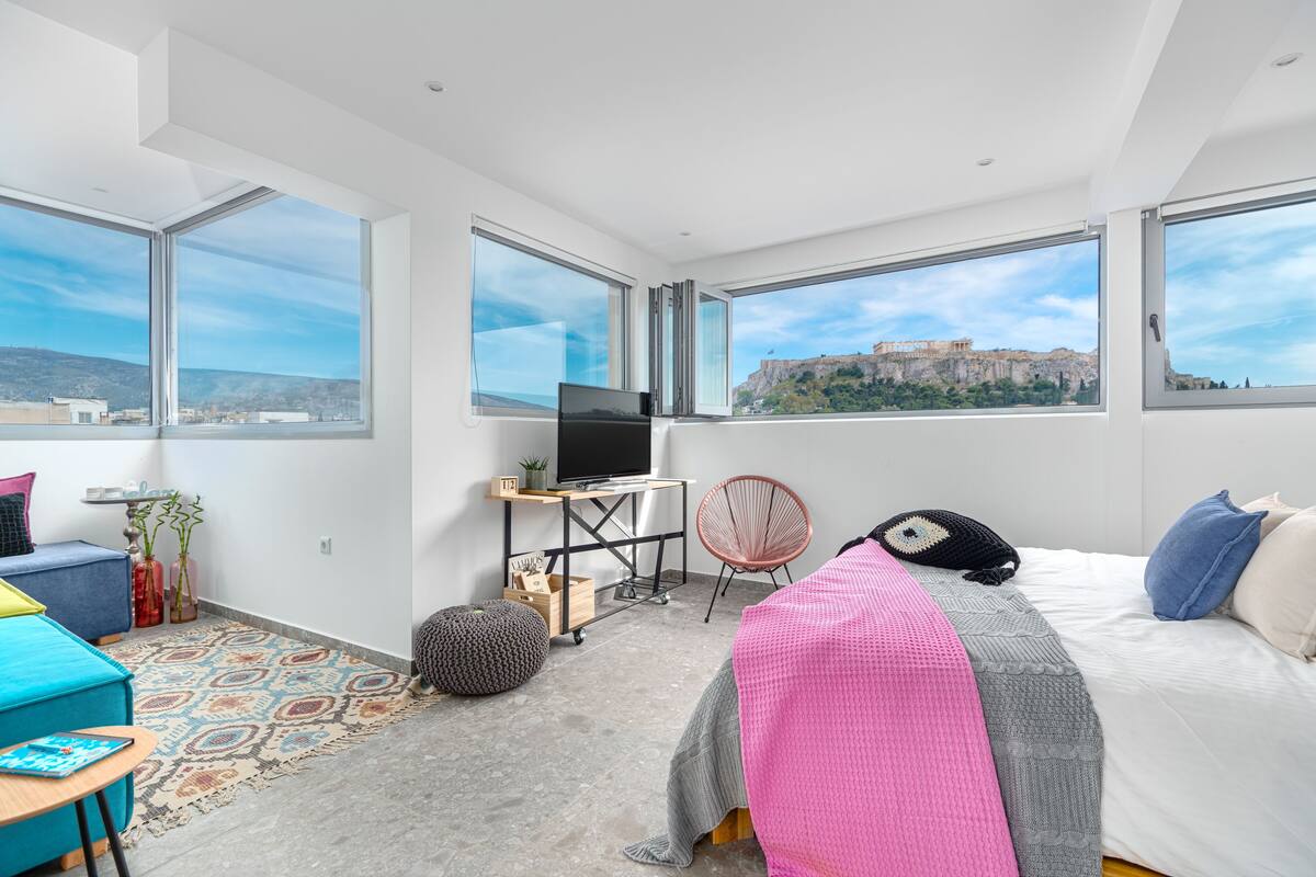 Una habitación de hotel moderna con ventanas amplias, una cama, un escritorio con televisor, una silla y vistas a un edificio histórico.