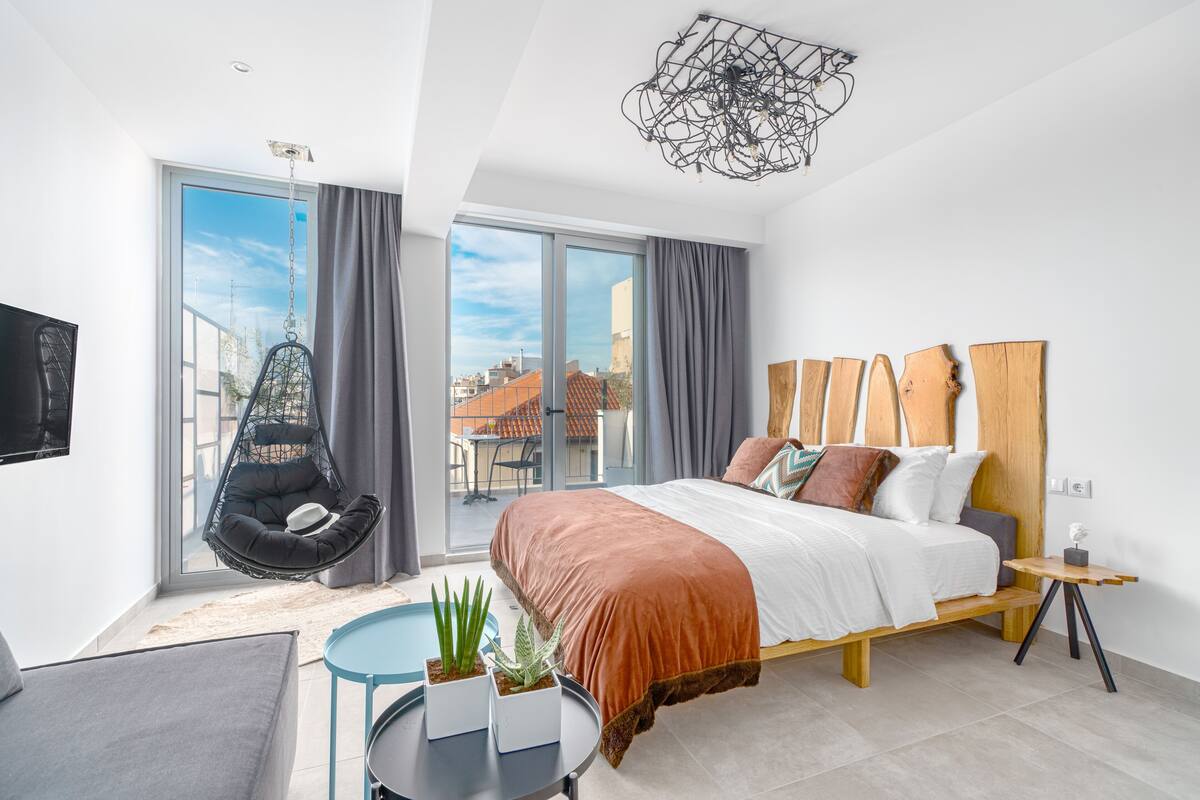 Un dormitorio moderno con una cama grande, una silla colgante, una mesita con plantas y vistas a la ciudad a través de amplios ventanales.