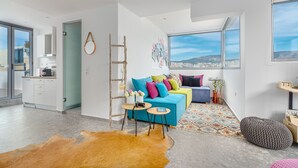 A modern living room with a blue sofa, colorful cushions, a wooden ladder, a small round table, and a large window offering a city view. - Mythos Luxury Suites (Athens)