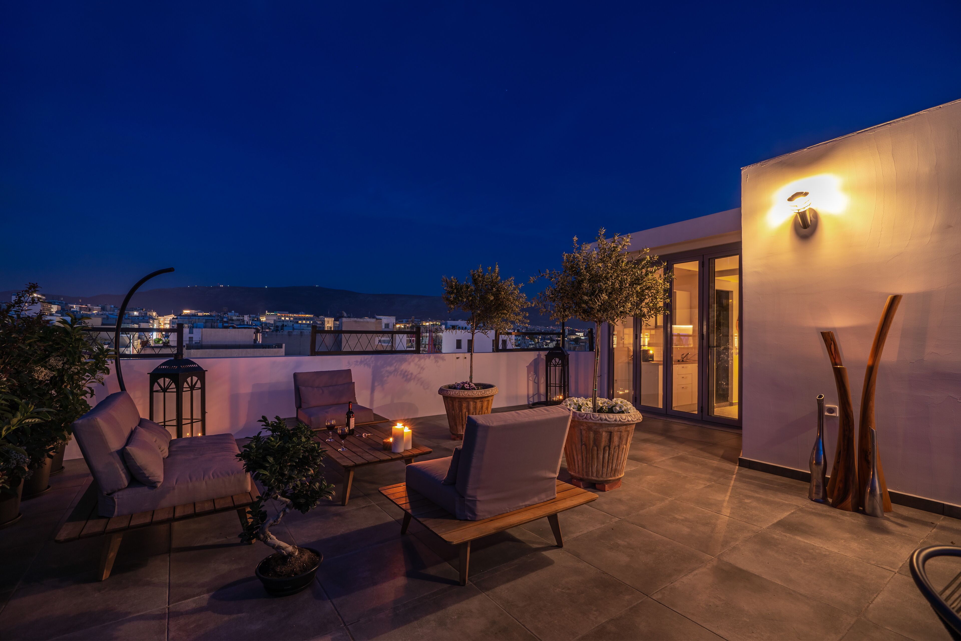 A rooftop terrace with outdoor seating, potted plants, and a city view at dusk.