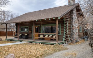 A log cabin with a porch, green-framed windows, and a stone chimney. - Scout's Rest in Cody - Beautiful Cozy Log Cabin that sleeps 8! (Cody)