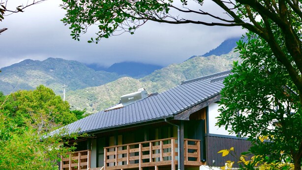A traditional Japanese building with a wooden deck, surrounded by lush greenery and mountains in the background. - Yakushima South Coast House (Yakushima)