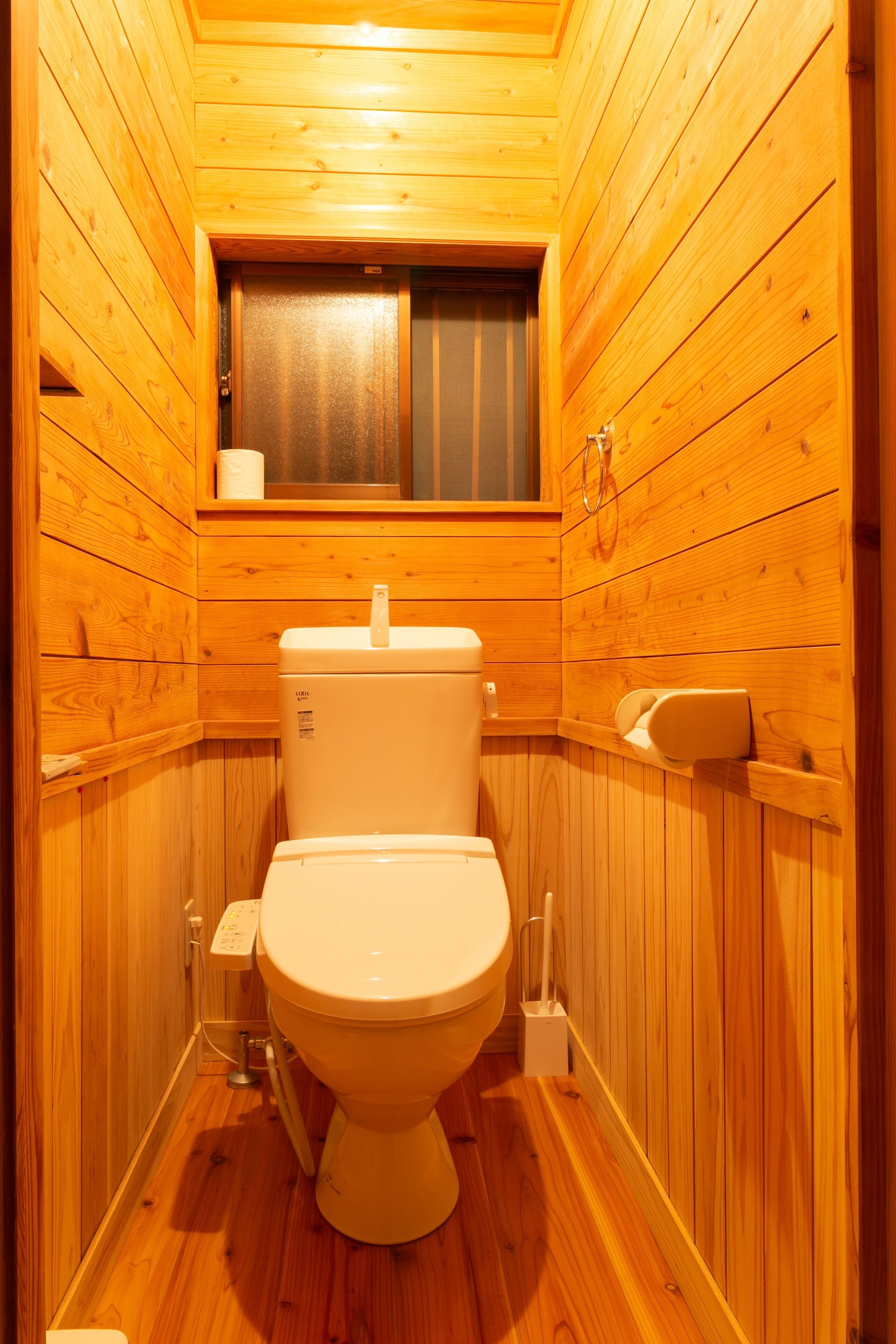 A small, wooden-paneled bathroom with a toilet, a window, and a shelf with a tissue box.