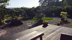 Une terrasse en bois avec un banc et des coussins, entourée d’un jardin agrémenté de plantes en pot et offrant une vue sur la mer.