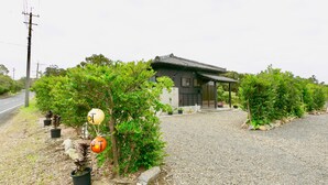 Une maison traditionnelle japonaise, avec un toit recouvert de tuiles, entourée d’une allée gravillonnée et de plantes en pot.