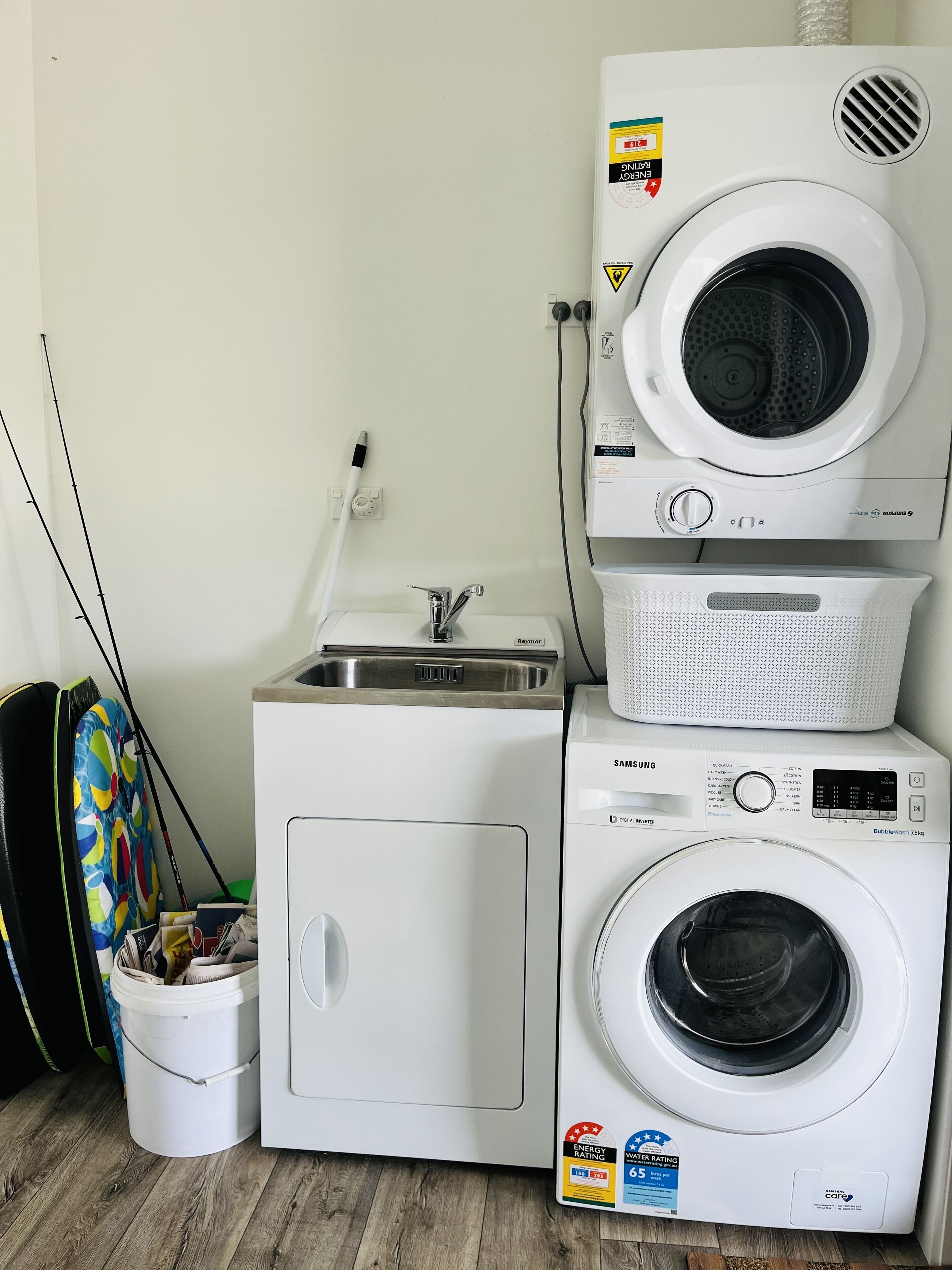 Een wasmachine en droger gestapeld, met een zichtbaar energielabel en waterverbruikslabel, een Raymar kraan en een witte emmer met spullen.