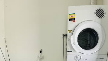 Een wasmachine en droger gestapeld, met een zichtbaar energielabel en waterverbruikslabel, een Raymar kraan en een witte emmer met spullen.