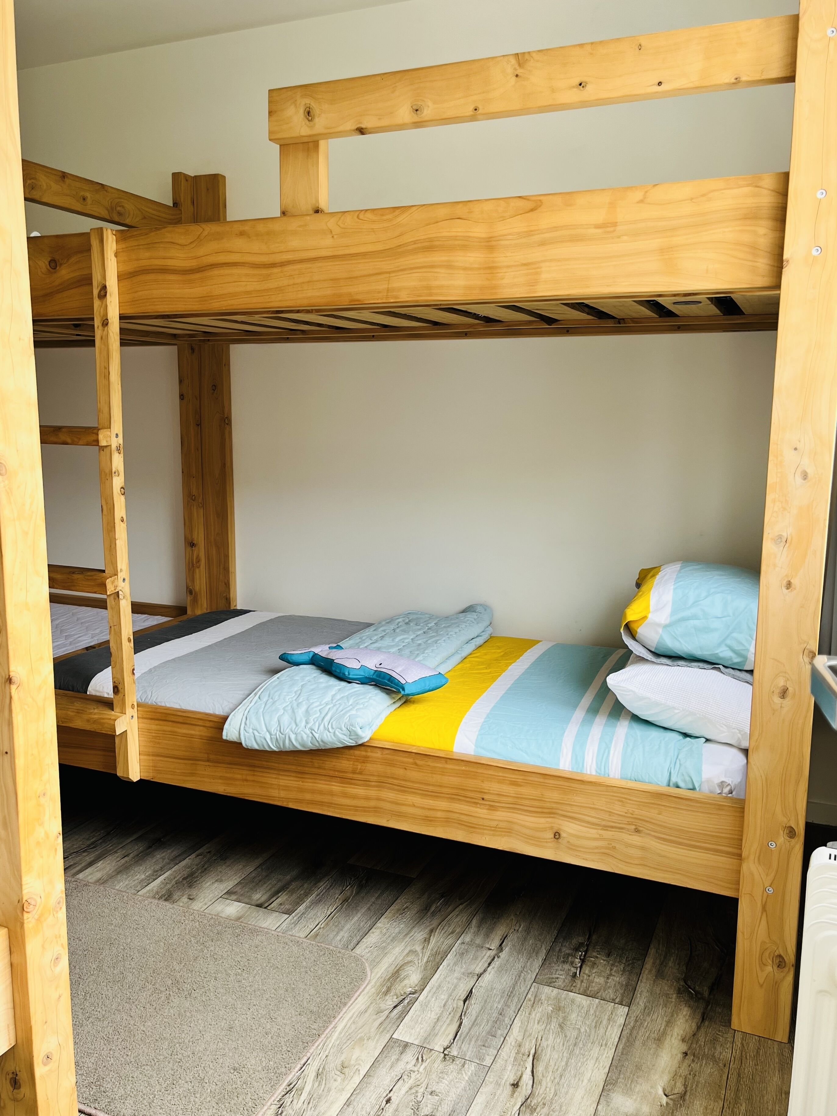A bunk bed with a wooden frame, a grey mat at the foot of the bed, and a grey wall in the background.