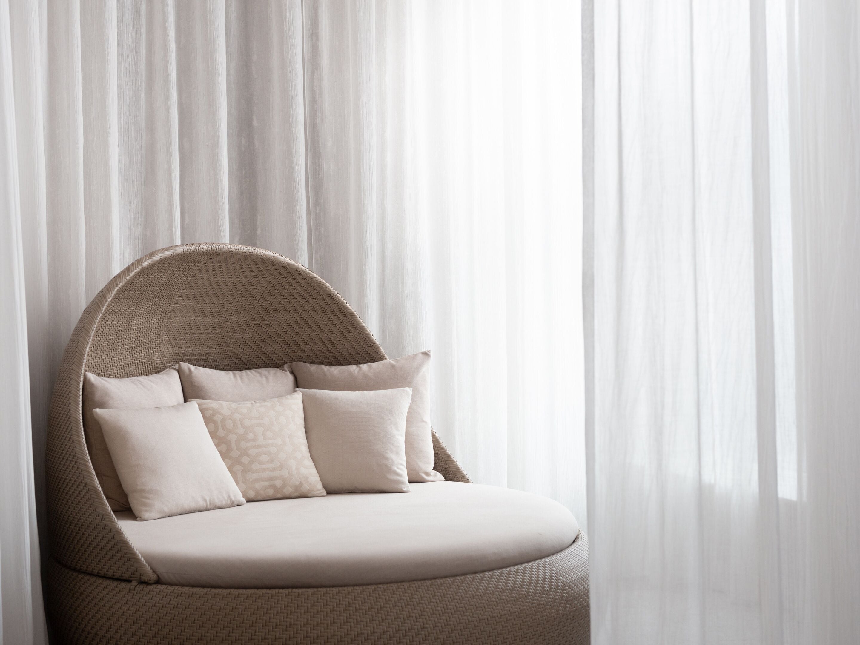 A wicker chair with a cushioned seat and backrest, positioned in front of sheer curtains.