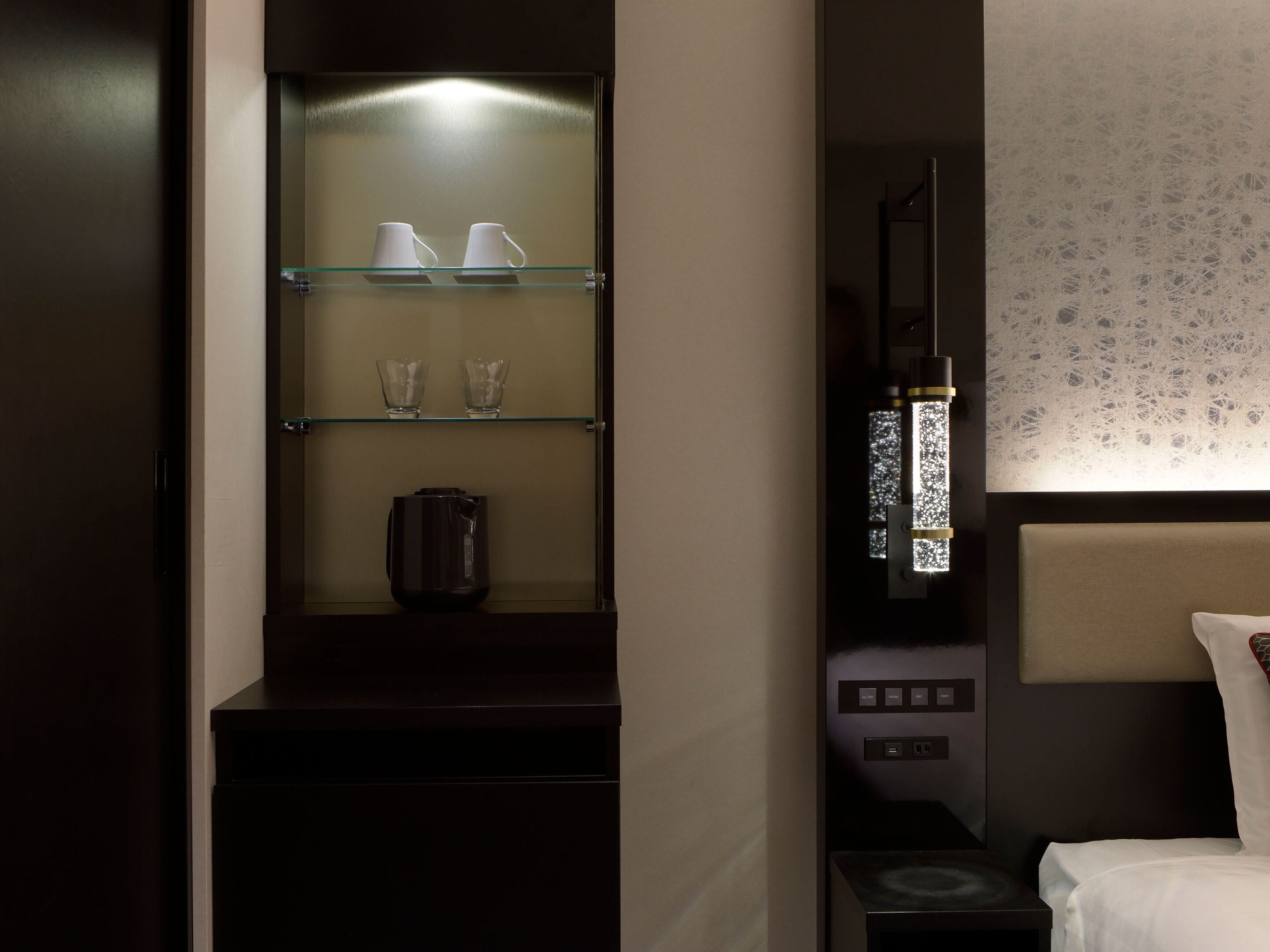 A hotel room with a dark wood cabinet containing a coffee maker and two cups. There is a bedside table with a lamp and a bed with a headboard.
