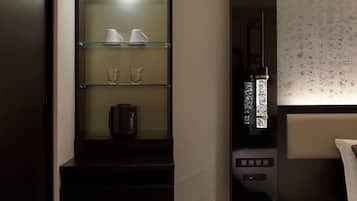 A hotel room with a dark wood cabinet containing a coffee maker and two cups. There is a bedside table with a lamp and a bed with a headboard.