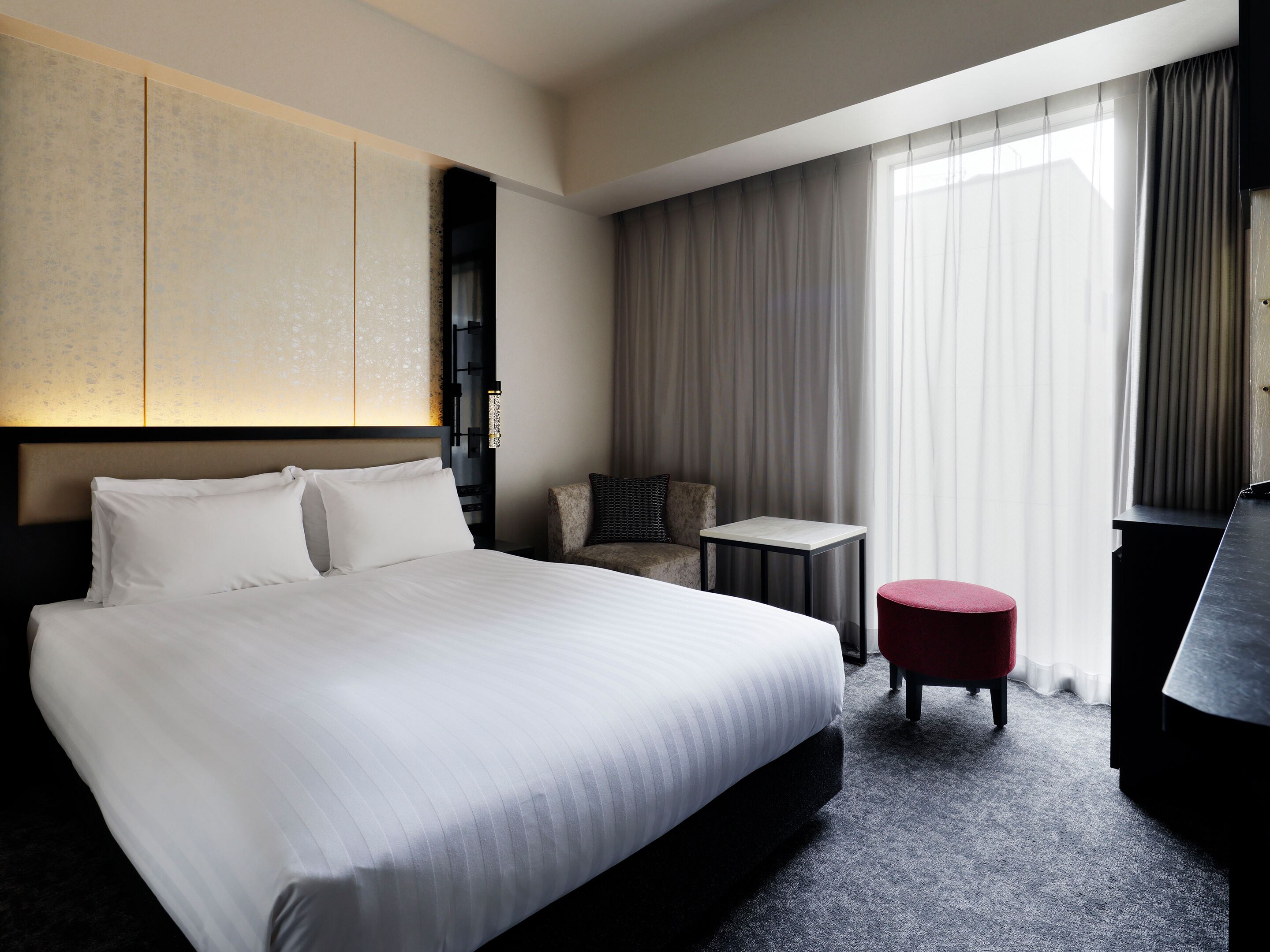 A hotel room with a large bed, a small table, a red ottoman, a sofa, and a large window with curtains.